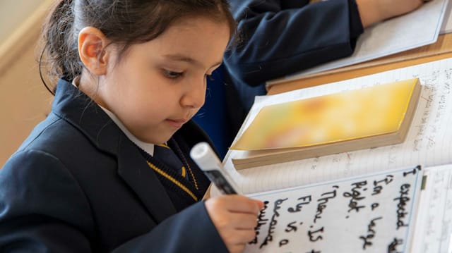 A pupil writes in a workbook at a classroom desk.