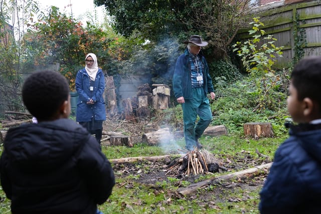 Pupils watch adults tend a small fire during a forest school activity.