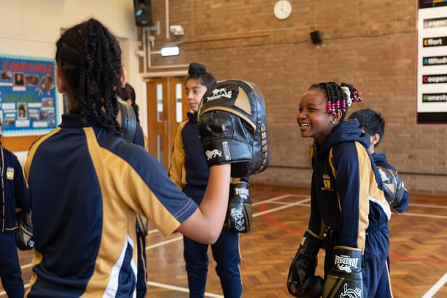 Pupils wearing boxing gloves practise pad work in the school hall.