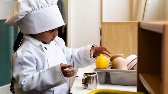 An early years pupil plays at a toy kitchen with a chef hat and utensils.