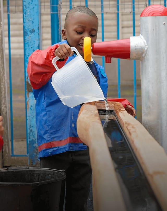 A young pupil pours water from a jug into an outdoor play trough.